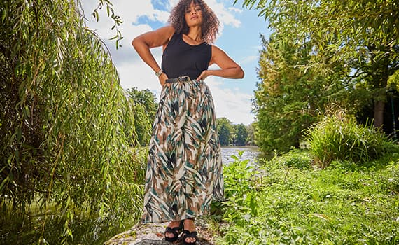 Frau im ärmellosen Top und langem Rock mit Blattmuster steht im Grünen am See